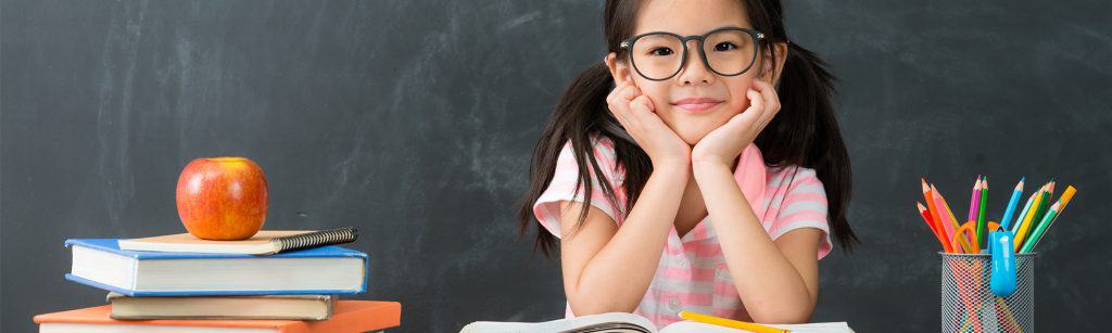 young girl sits in front of blackboard at desk, smiling, with books and pencils and more around her