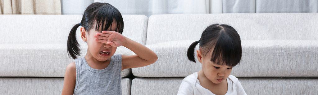 Two young girls at home have sibling rivalry, sit in front of couch, one playing with toys while the other cries and is upset