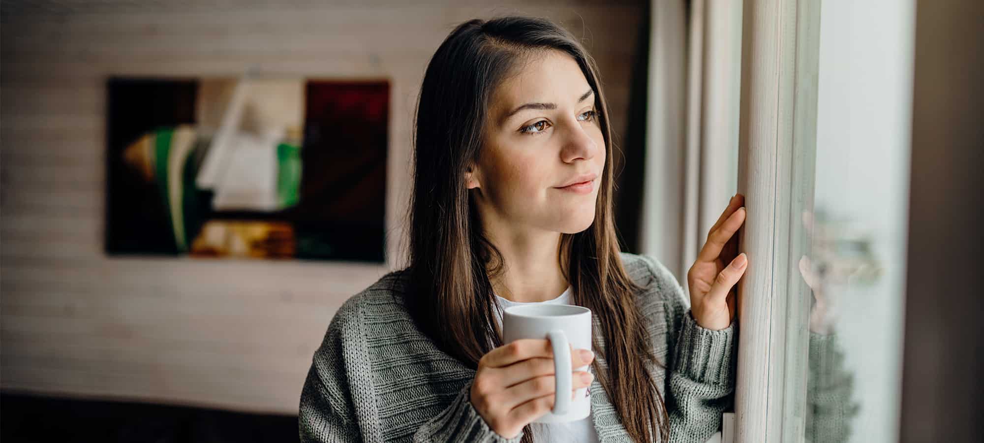 Woman looks longing out of window while holding cup